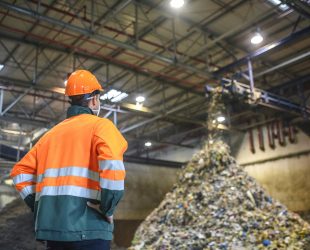 Low angle rear view of young male worker in helmet, pollution mask, and reflective clothing observing waste falling from conveyor belt onto pile at facility.