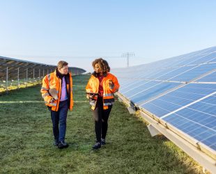 Male and female engineers working at solar power plant. Two technicians in reflective clothing walking between rows of photovoltaic panels at solar farm.