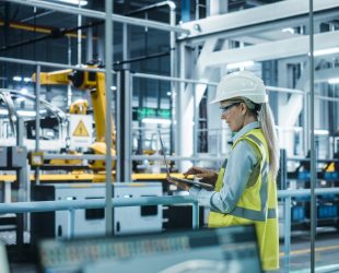 Car Factory: Female Automotive Engineer Wearing Hard Hat, Standing, Using Laptop. Monitoring, Control, Equipment Production. Automated Robot Arm Assembly Line Manufacturing Electric Vehicles.