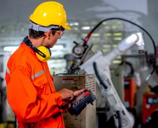 Factory worker man hold controller to control metal welding robotic machine and work in workplace area. Industrial and technology support system help in working of employee concept.