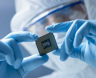 In Ultra Modern Electronic Manufacturing Factory Design Engineer in Sterile Coverall Holds Microchip with Gloves and Examines it.