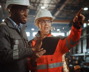 Two Heavy Industry Engineers Stand in Steel Metal Manufacturing Factory, Use Digital Tablet Computer and Have a Discussion. Black African American Industrial Specialist Talk to Female Technician.