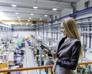 Horizontal color image of blond manager working with digital tablet in large futuristic factory. Blond woman standing on top of a balcony. Focus on businesswoman, futuristic machines in background.