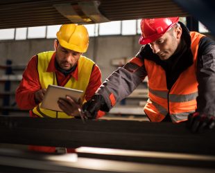 Two manual workers measuring metal in a factory, while one of them is using digital tablet. Focus is on man with yellow hardhat.